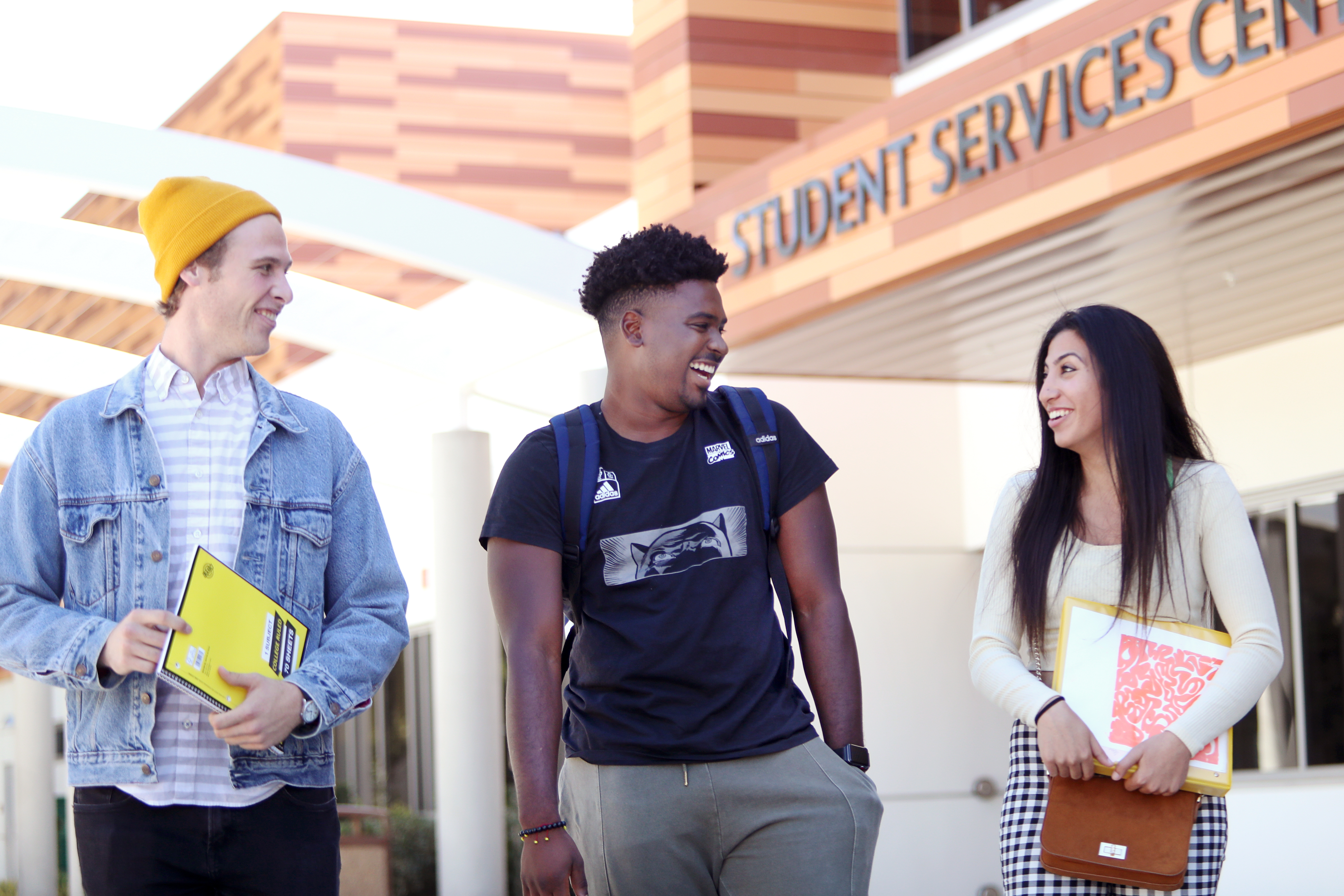 Three college students smiling and walking together outside the Student Services Center, holding notebooks and looking engaged.