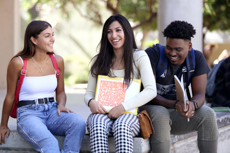 Three college students sitting outdoors on a campus ledge, smiling and chatting while holding notebooks and wearing backpacks.