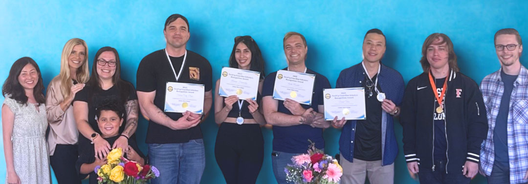 Group of graduates holding certificates and medals while posing against a blue backdrop.