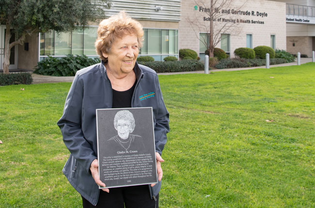 Cathy Crown standing on a campus lawn holding a commemorative plaque, with a school building in the background.