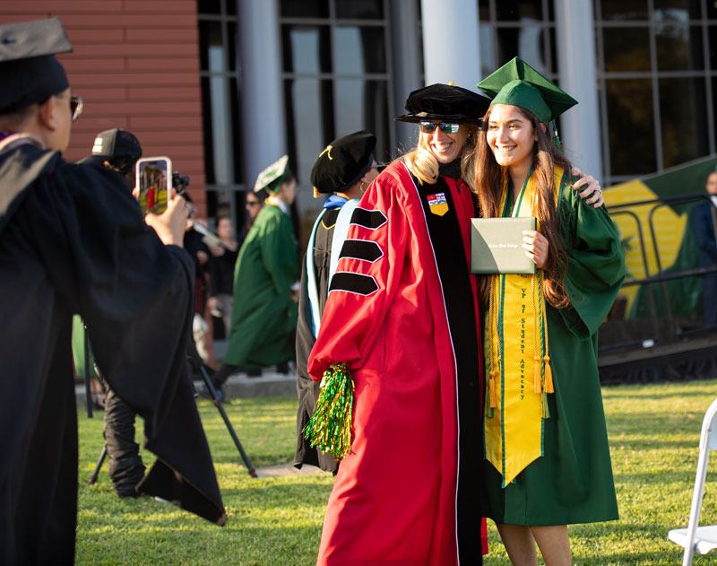 Graduate in green cap and gown holds a diploma and smiles while posing with a faculty member in academic regalia at an outdoor commencement.