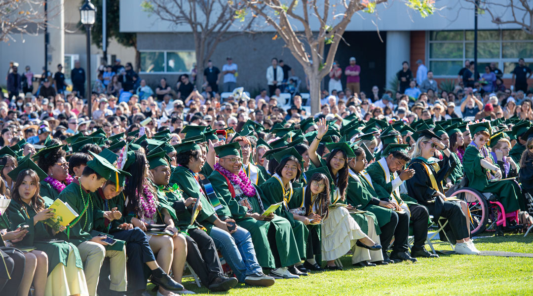 Outdoor graduation ceremony with rows of students in green caps and gowns seated on a lawn, with a large crowd of attendees behind them.