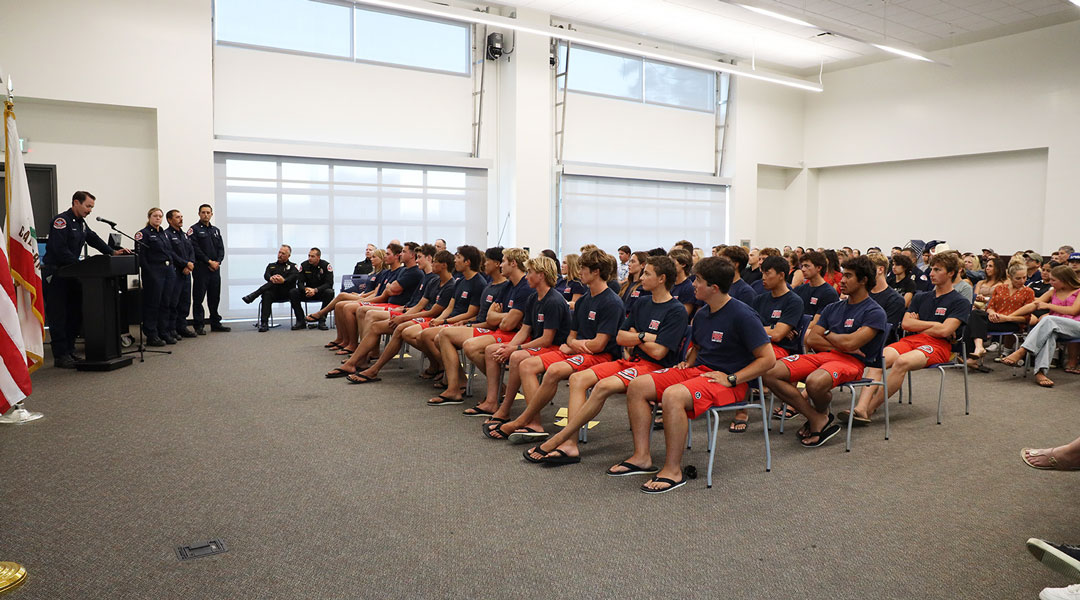 Large group of uniformed trainees seated in rows listening to a speaker at a podium in a bright classroom, with several officials standing at the front.